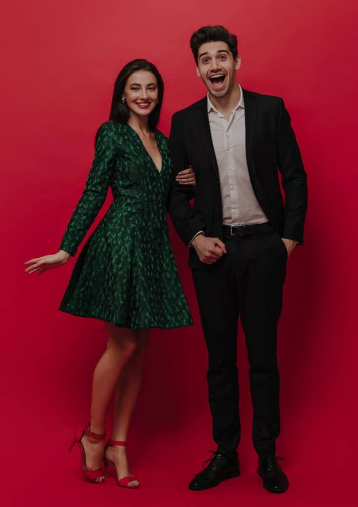 nedagorgeous young girl with green midi dress high heels smiling holding hand her excited boyfriend white shirt black suit red plain background scaled e1666532142943